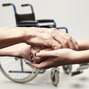 Hands of an elderly man holding the hand of a younger woman in front of a wheel chair.